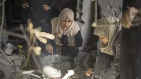 Two women in hijabs cooks over a makeshift fire in a damaged building in Gaza.