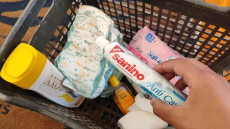  Hand holding toothpaste over shopping basket. Basket contains hygiene products including diapers, toiletries, and sanitary items.