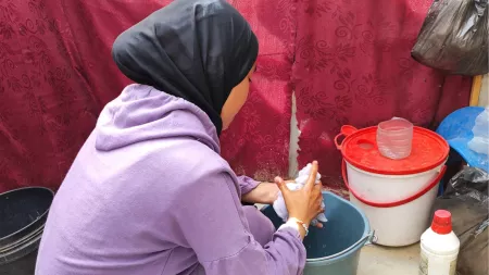 Woman in headscarf reaches for hygiene products on shelf in her makeshift tent.