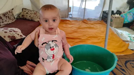 A woman holding her baby in a makeshift tent. There's a green bucket on their side.