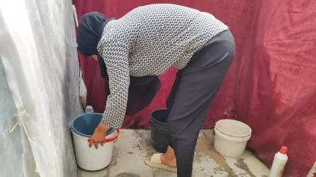 A woman in black headscarf, white patterned shirt and black pants bend over to pick a white bucket with red handles. She is in a red makeshift tent.