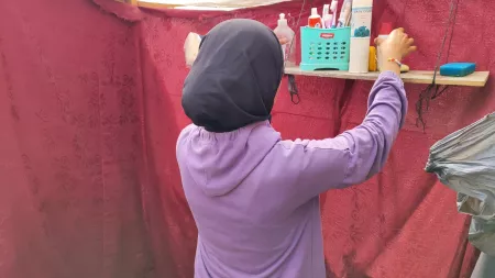 Girl in headscarf reaches for hygiene products on shelf in a red makeshift tent. 