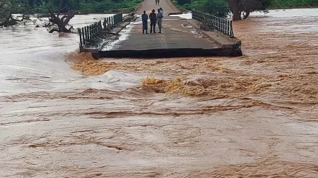 Three people stand on a damaged bridge over a raging river, with floodwaters rushing below and surrounding trees partially submerged.