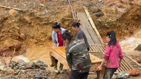 Four women carry a wooden table across a muddy, rocky area near a stream, after a flood.