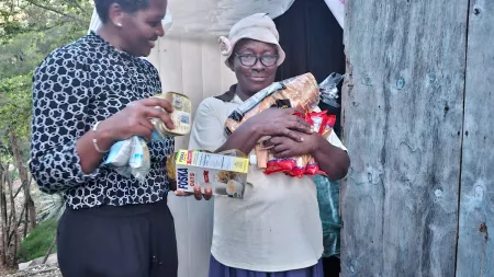 Two women stand outside a makeshift shelter. One woman in a patterned top holds food items, while an older woman in a hat and glasses carries groceries and packaged goods.