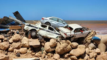 A pile of wrecked cars and debris stacked on a rocky shoreline, remnants of destruction caused by severe flooding in Libya.