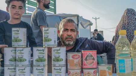 A man and a boy stand behind a table displaying canned food and cooking oil at an outdoor market or aid distribution point, with people visible in the background.