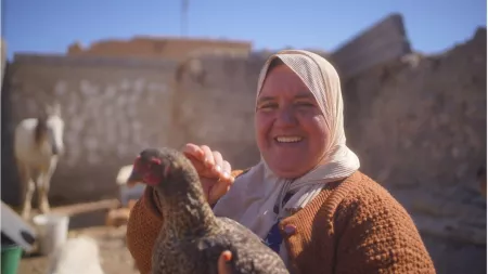 A smiling woman in a headscarf holds a chicken in her arms in an outdoor area with a stone wall in the background.