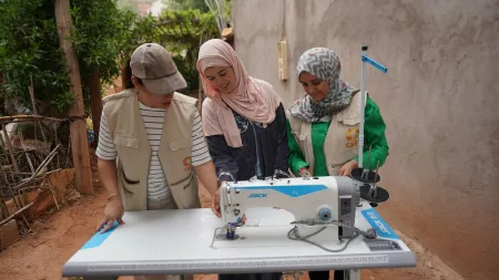 Three women, including two CARE Maroc team members and a smiling woman in a headscarf, gather around a sewing machine outdoors. 