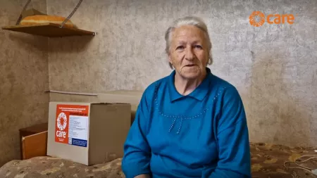 Elderly woman in blue sweater sits next to CARE aid box in sparse room.