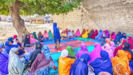 Circle of women wearing colorful heardscarfs around one woman standing. Group is outdoors.