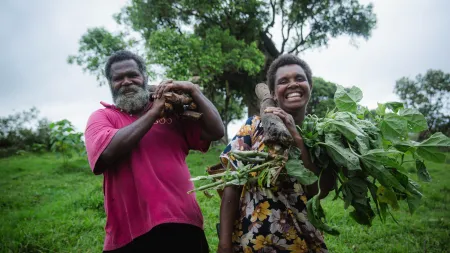 Man and woman smiling to the camera while carrying wood and leave trees in a green area