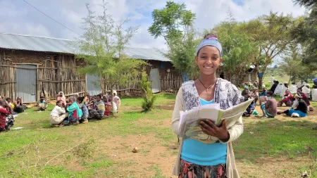 Girl standing in outdoor space, holding book while smiling to the camera