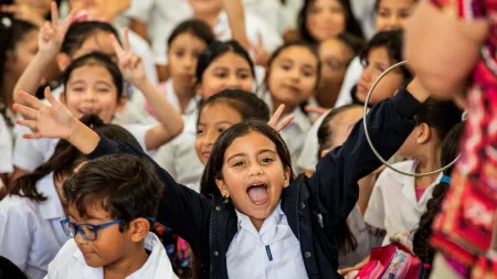 Large group of school children sitting down while signaling to the camera. One girl at the centre is with wide lifted arms and open mouth in joy.
