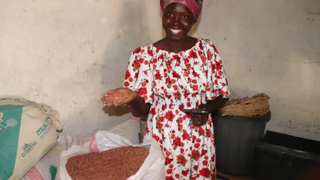 Woman withwearing white and red flowe dress smiling to the camera while showing a handful of grains, in front of a bag of the same grains