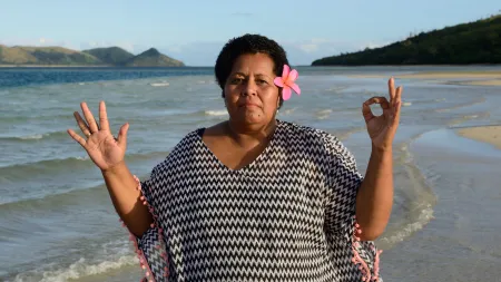 Woman at the beach wearing black and white patterned shirt, flower on her short hair, and both hands up - one with an okay sign.