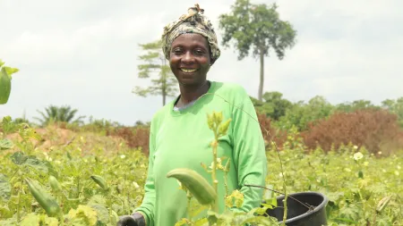 Woman wearing green shirt and head scarf. She's standing in a plantation and smiling to the camera.