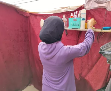 Girl in headscarf reaches for hygiene products on shelf in a red makeshift tent. 