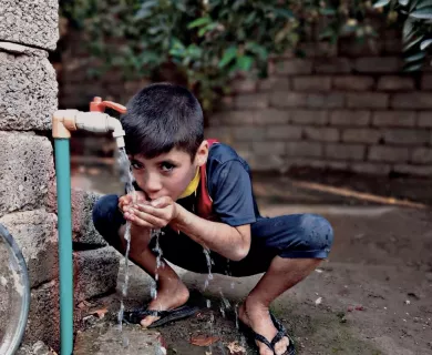 Young boy crouches to drink water directly from outdoor tap, cupping hands to catch flow. Brick wall and plants in background.