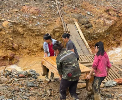 Four women carry a wooden table across a muddy, rocky area near a stream, after a flood.