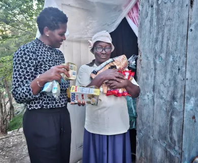 Two women stand outside a makeshift shelter. One woman in a patterned top holds food items, while an older woman in a hat and glasses carries groceries and packaged goods.