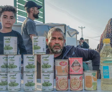 A man and a boy stand behind a table displaying canned food and cooking oil at an outdoor market or aid distribution point, with people visible in the background.