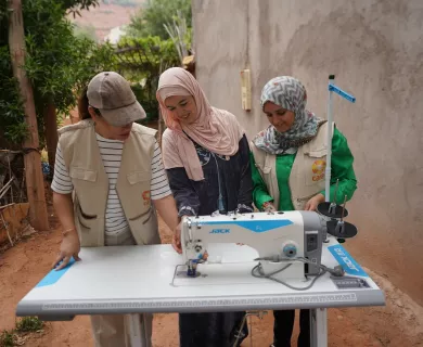 Three women, including two CARE Maroc team members and a smiling woman in a headscarf, gather around a sewing machine outdoors. 