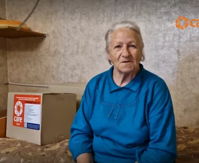 Elderly woman in blue sweater sits next to CARE aid box in sparse room.