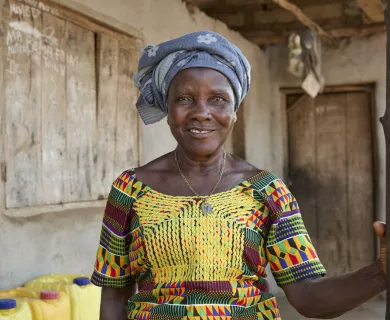 Woman wearing patterned dress and headscarf standing in wooden house porch, smiling to the camer