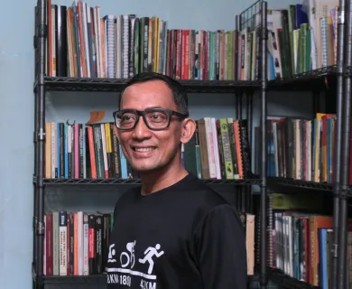 Man standing in front of bookshelf, looking to the left. The man has short hair, wears glasses and is gently smiling.